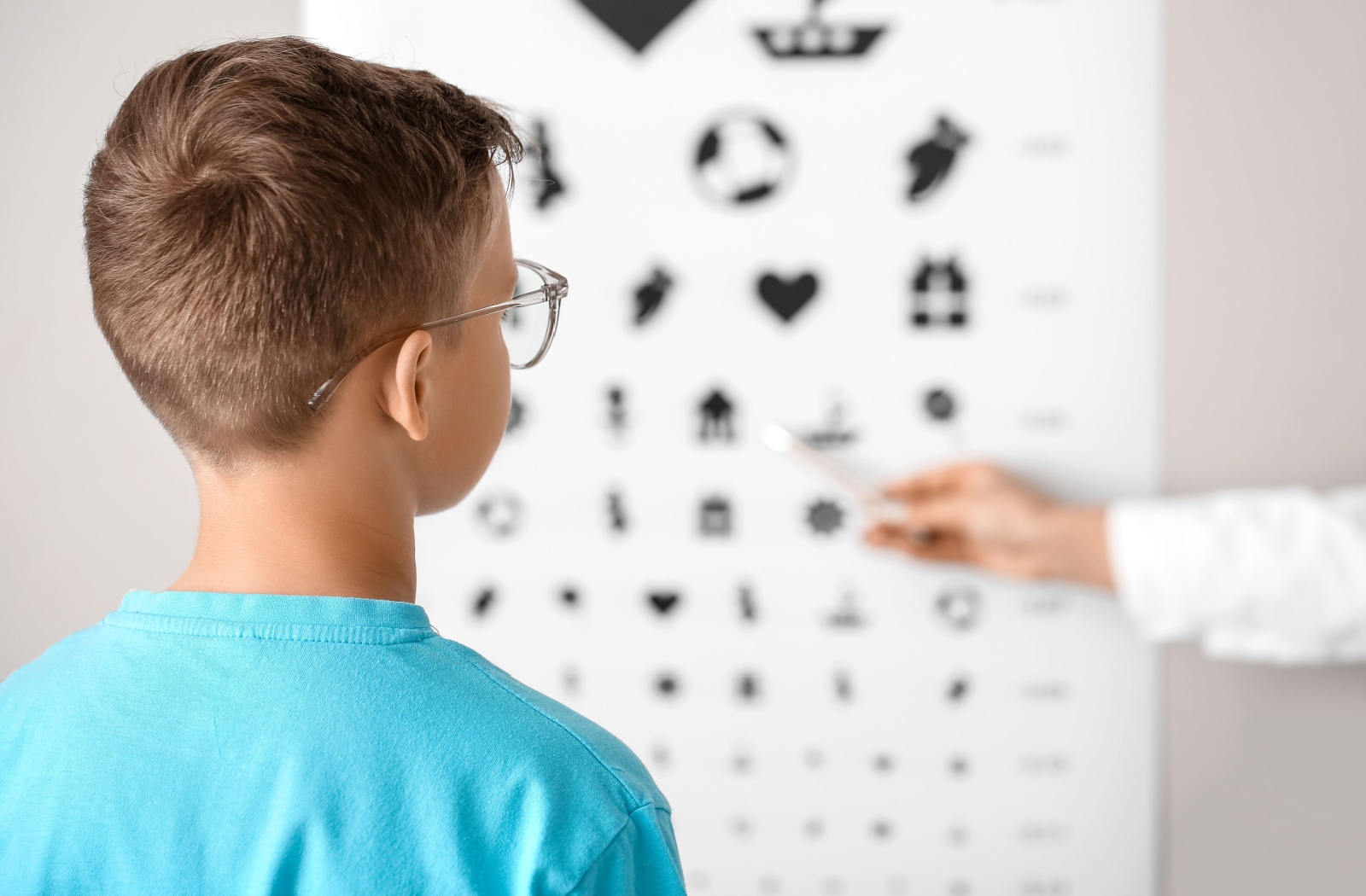 An optometrist points at a symbol on an eye chart while measuring a young boy's vision