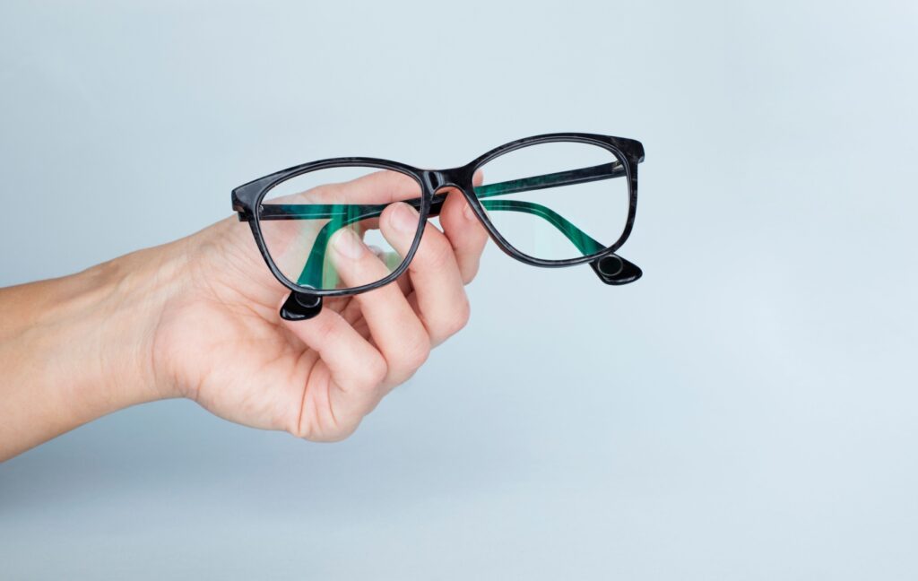 a hand holds out a pair of black-rimmed glasses in front of a pale blue background.